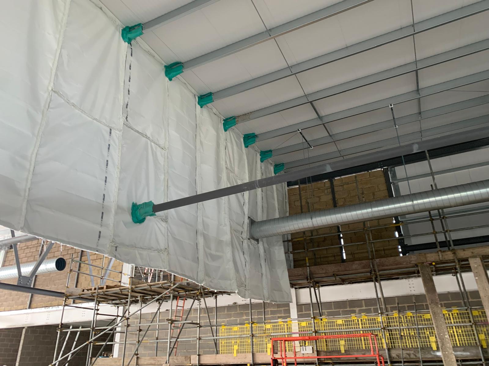 Passive fire barrier hung from the roof steelwork of a commercial building, secured with green-wrapped fixings at each bracket