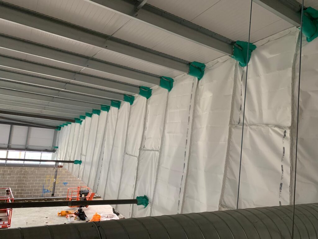 Long run of white passive fire barrier fixed along roof steelwork, with green-wrapped fixings at each support